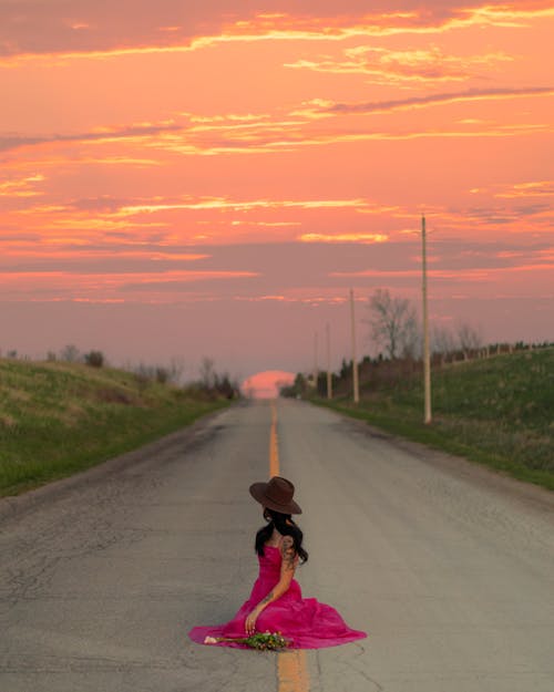 Young Woman in a Pink Dress and a Hat Sitting in the Middle of the Road at Sunset 