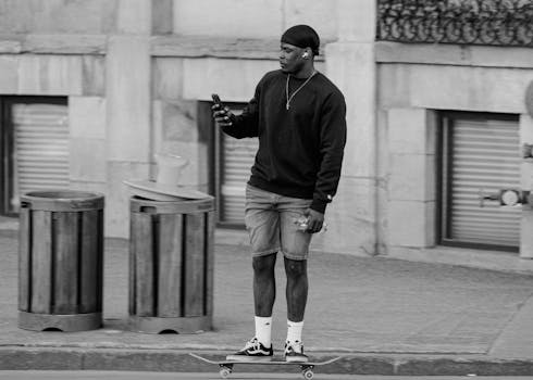 A man skateboards on a city sidewalk while using a cellphone, blending technology with urban lifestyle.
