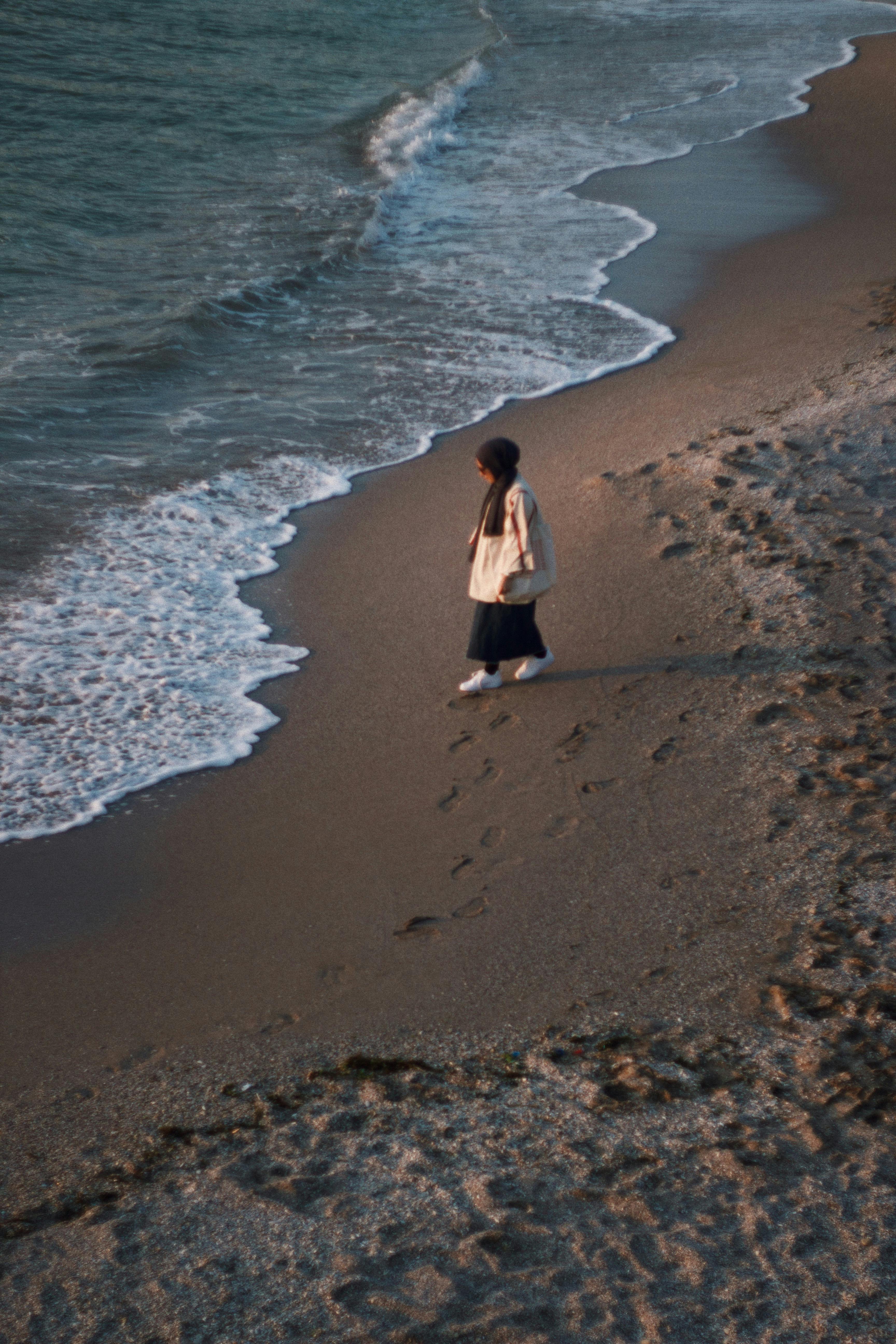 A woman in a hijab walks along a sandy beach at sunset, leaving footprints in the sand.