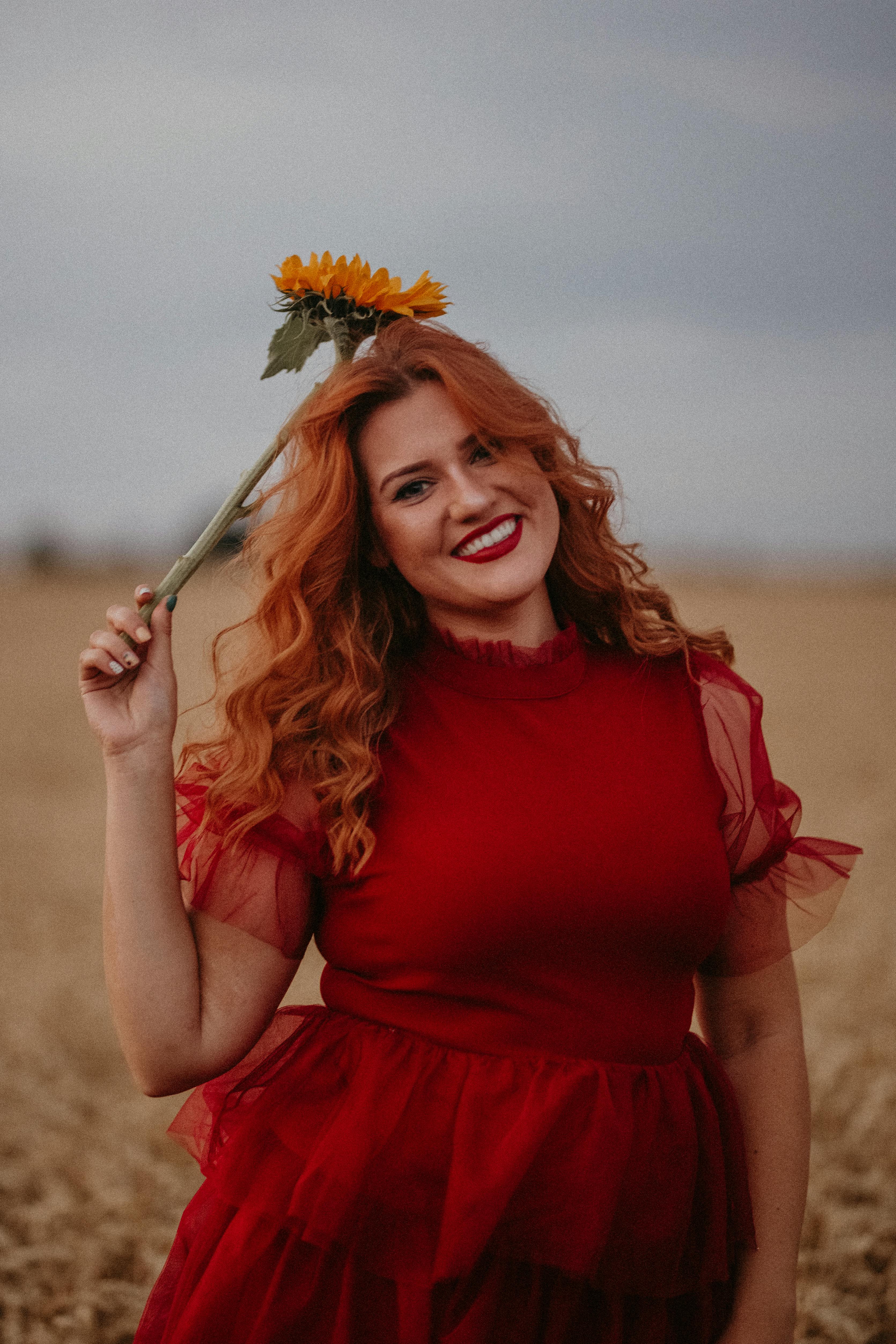 Woman in a red dress smiling while holding a sunflower in a wheat field in Colchester, England.
