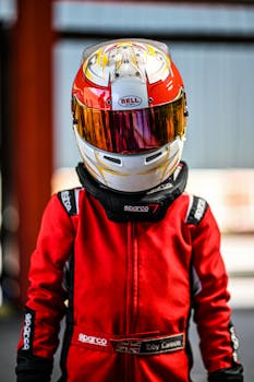 A child in a red racing suit and helmet stands ready for go-karting on a bright day.
