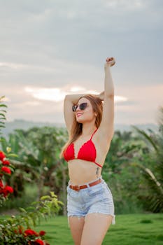 Stylish woman in red bikini top and denim shorts poses cheerfully outdoors during summer.