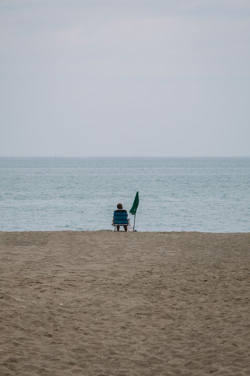 Free A tranquil view of a person sitting on a beach in Málaga, Spain, with a beach umbrella and the sea in the background. Stock Photo