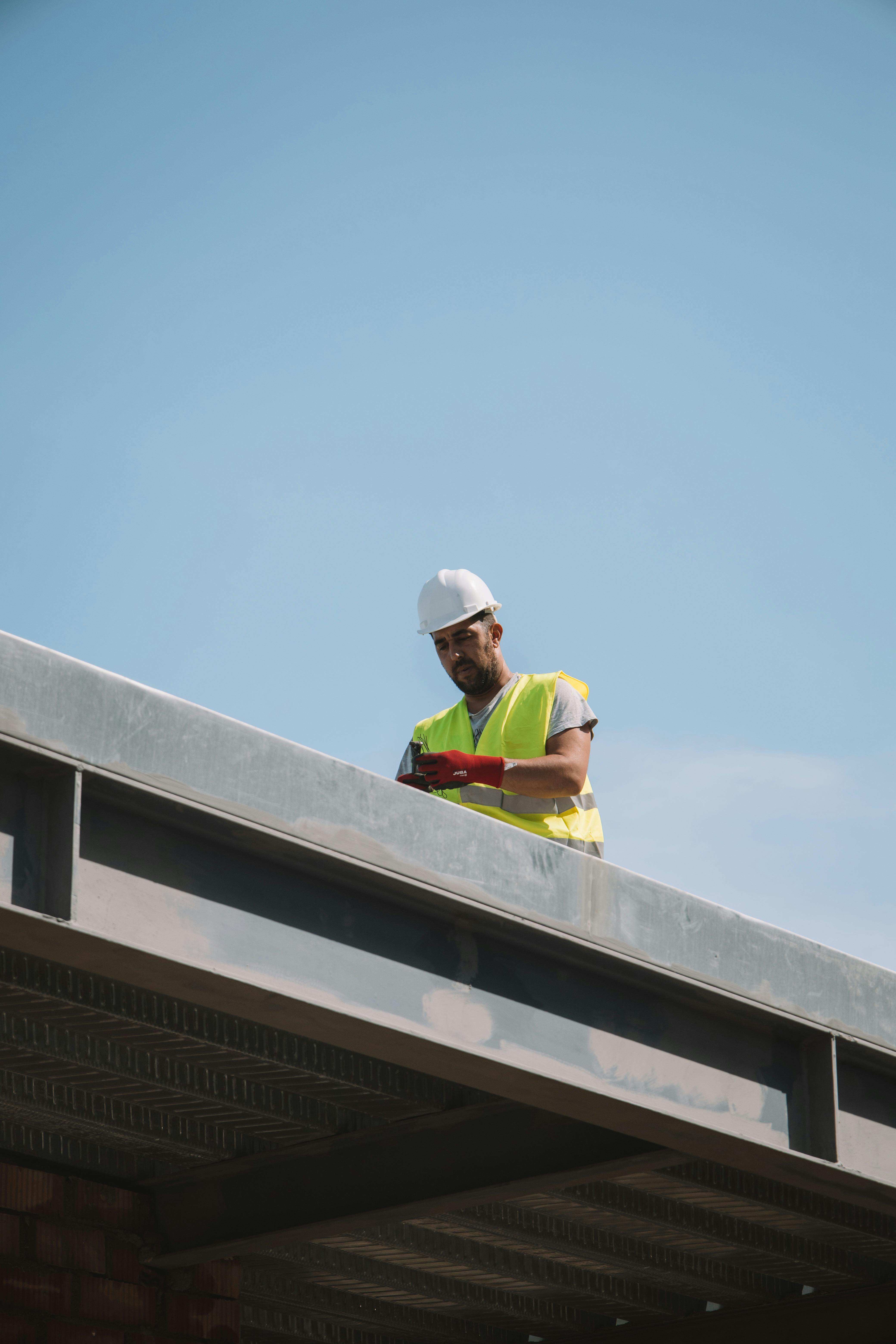 A roofer in safety gear standing on a commercial rooftop, viewed against a clear sky.