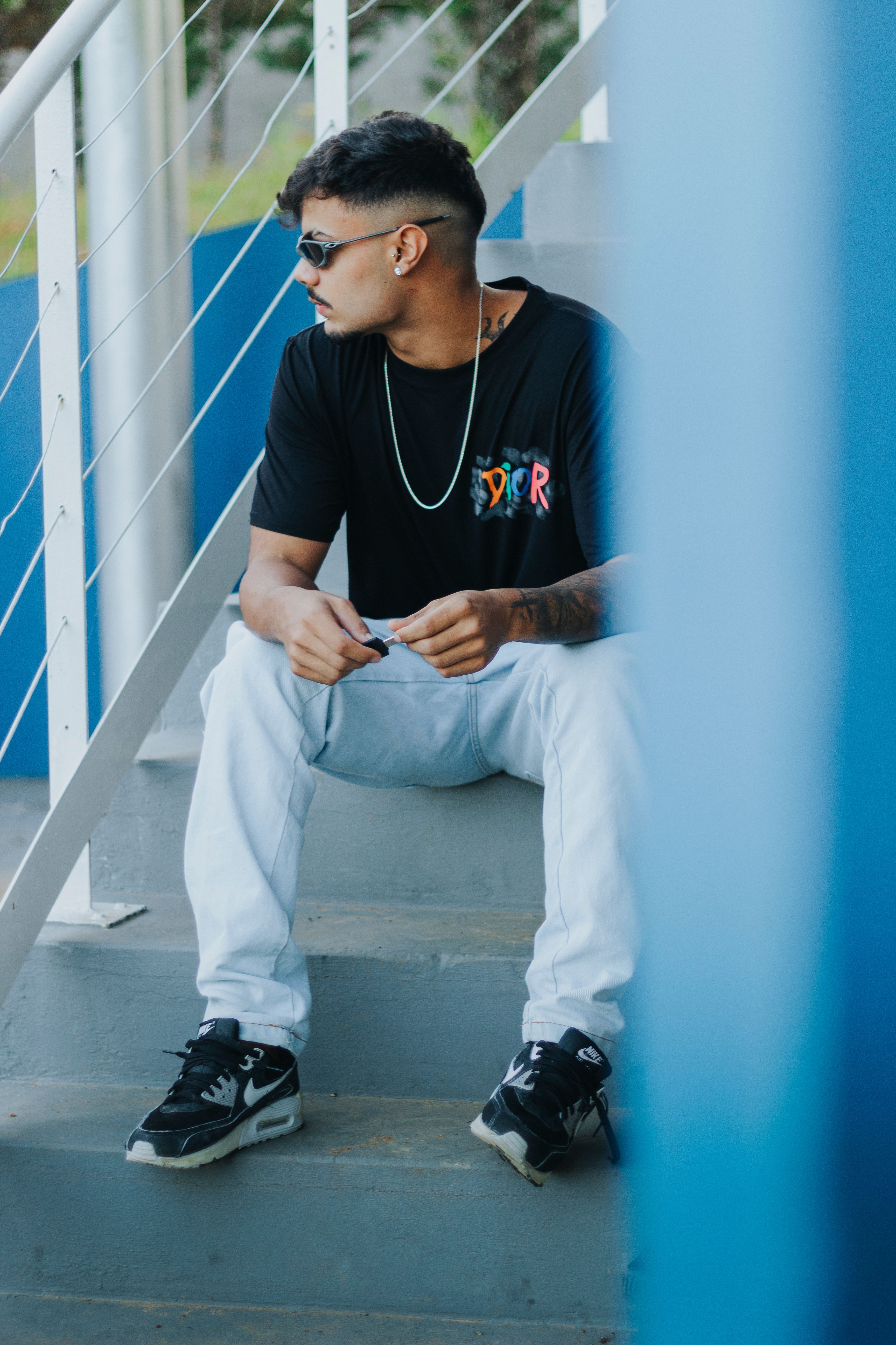 A young man in casual fashion sits on outdoor stairs, wearing sunglasses and a black t-shirt.