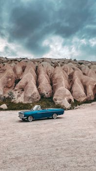 A classic blue convertible parked outdoors with striking rock formations and dramatic skies.