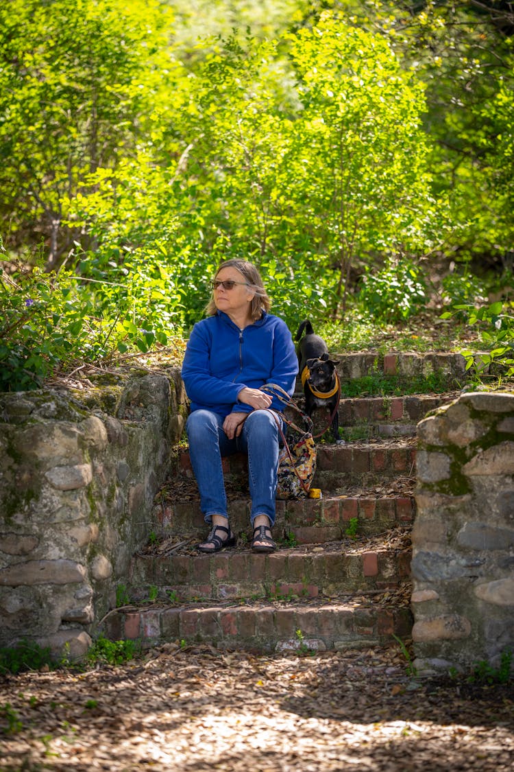 Woman With A Small Black Dog On A Walk Sitting On The Stairs In The Park