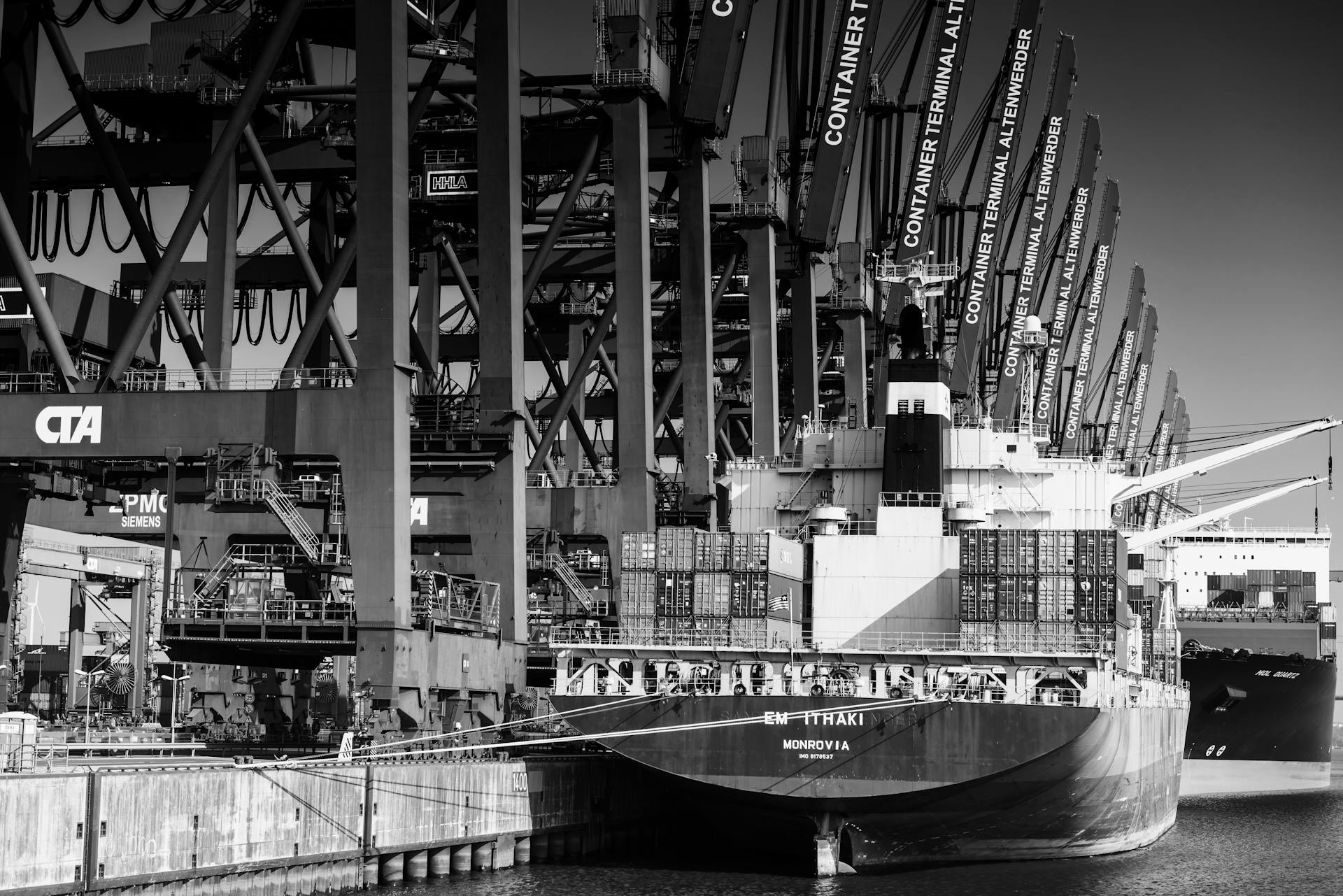 Monochrome photo of a large container ship docked at a bustling industrial port.