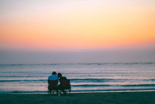 Free Couple enjoying a serene sunset while sitting on the beach, facing the sea. Stock Photo