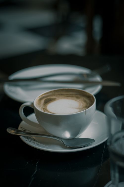 Free Close-up of a coffee cup with artistic froth, on a dark table setting with spoon and saucer. Stock Photo