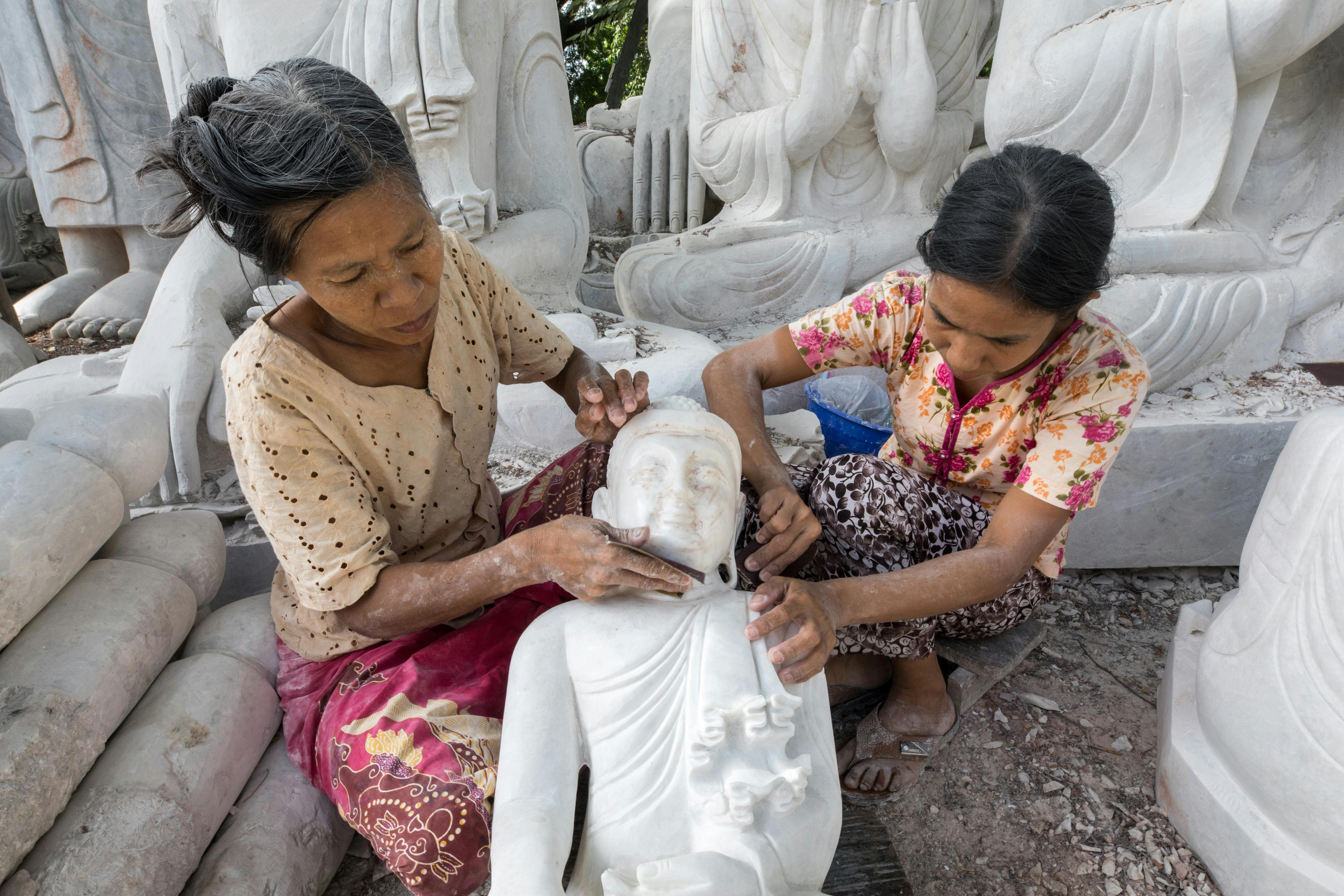 Two women skillfully crafting marble Buddha sculptures, showcasing traditional artisan techniques.