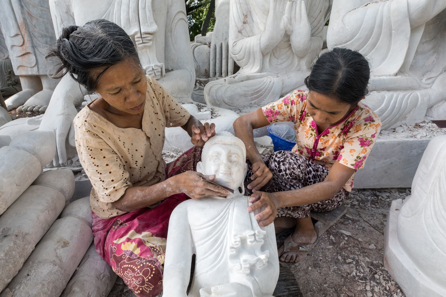 Two women skillfully crafting marble Buddha sculptures, showcasing traditional artisan techniques.