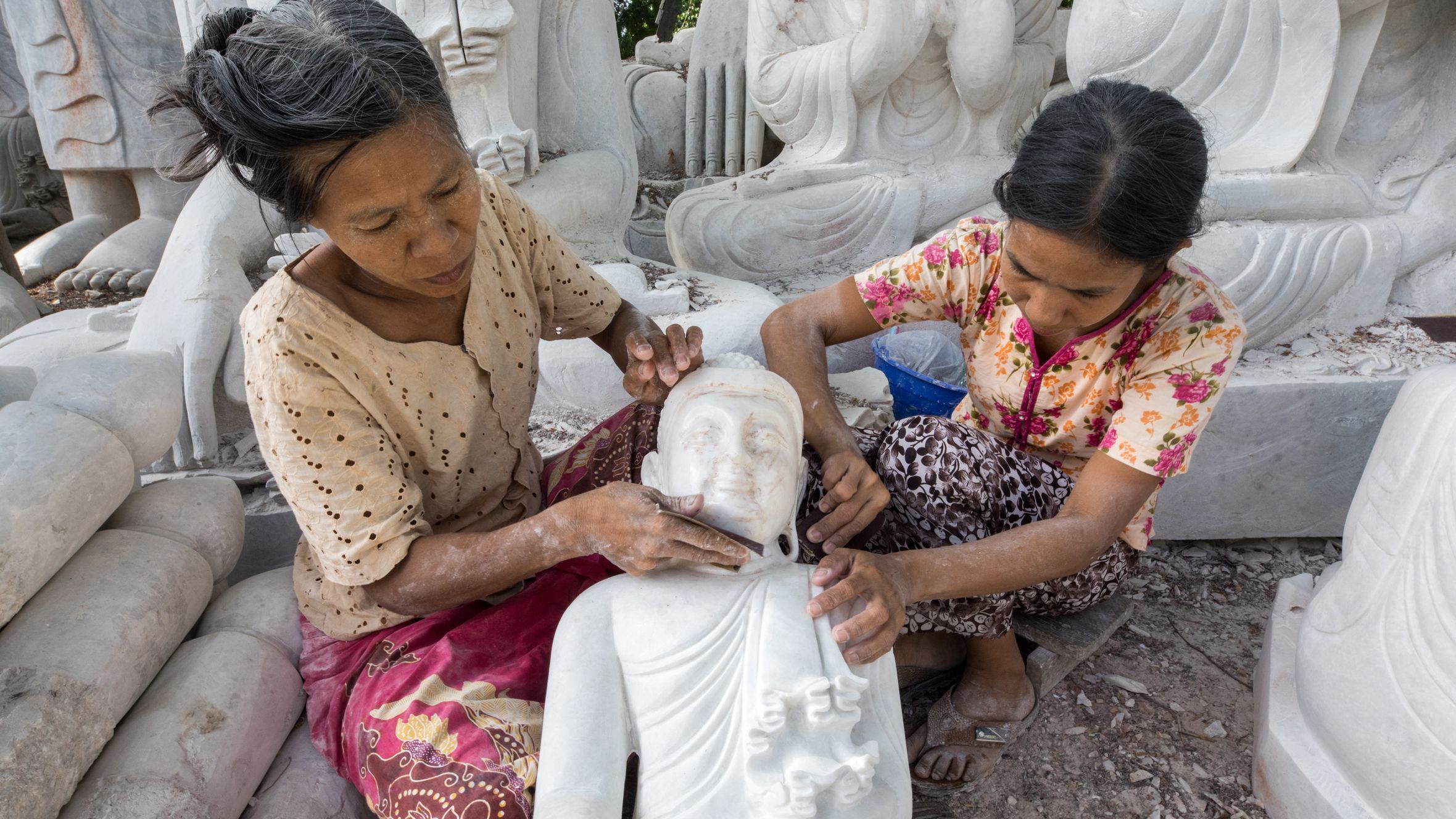 Two women skillfully crafting marble Buddha sculptures, showcasing traditional artisan techniques.