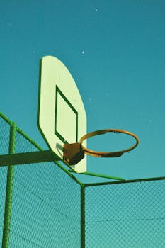 A sunlit basketball hoop against a clear blue sky with urban fencing.