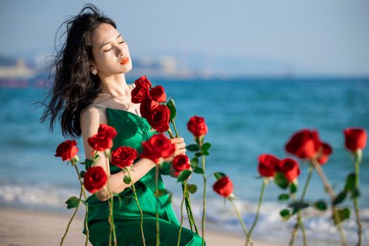 Woman in a green dress holding red roses by the sea, eyes closed, enjoying the serene beach atmosphere.
