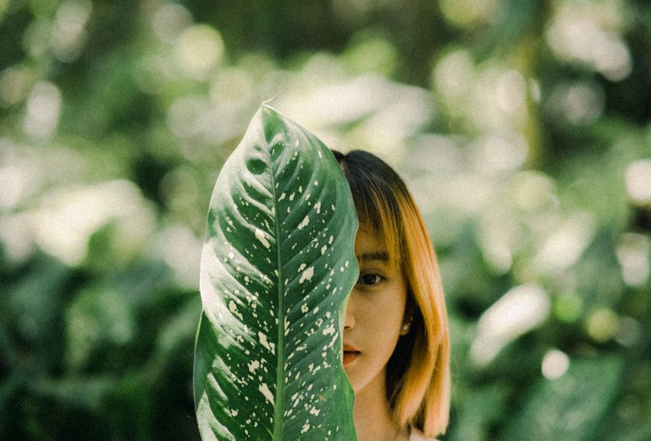 Young woman partially obscured by a large leaf in a lush forest setting.