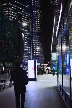 A man walks along a bustling city sidewalk illuminated by skyscraper lights at night.