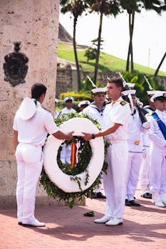 Navy officers in white uniforms holding a commemorative wreath at an outdoor ceremony.