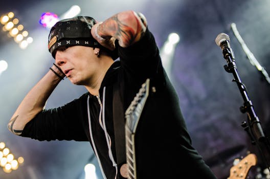 A tattooed rock musician performing energetically with a guitar and microphone onstage under bright lights.