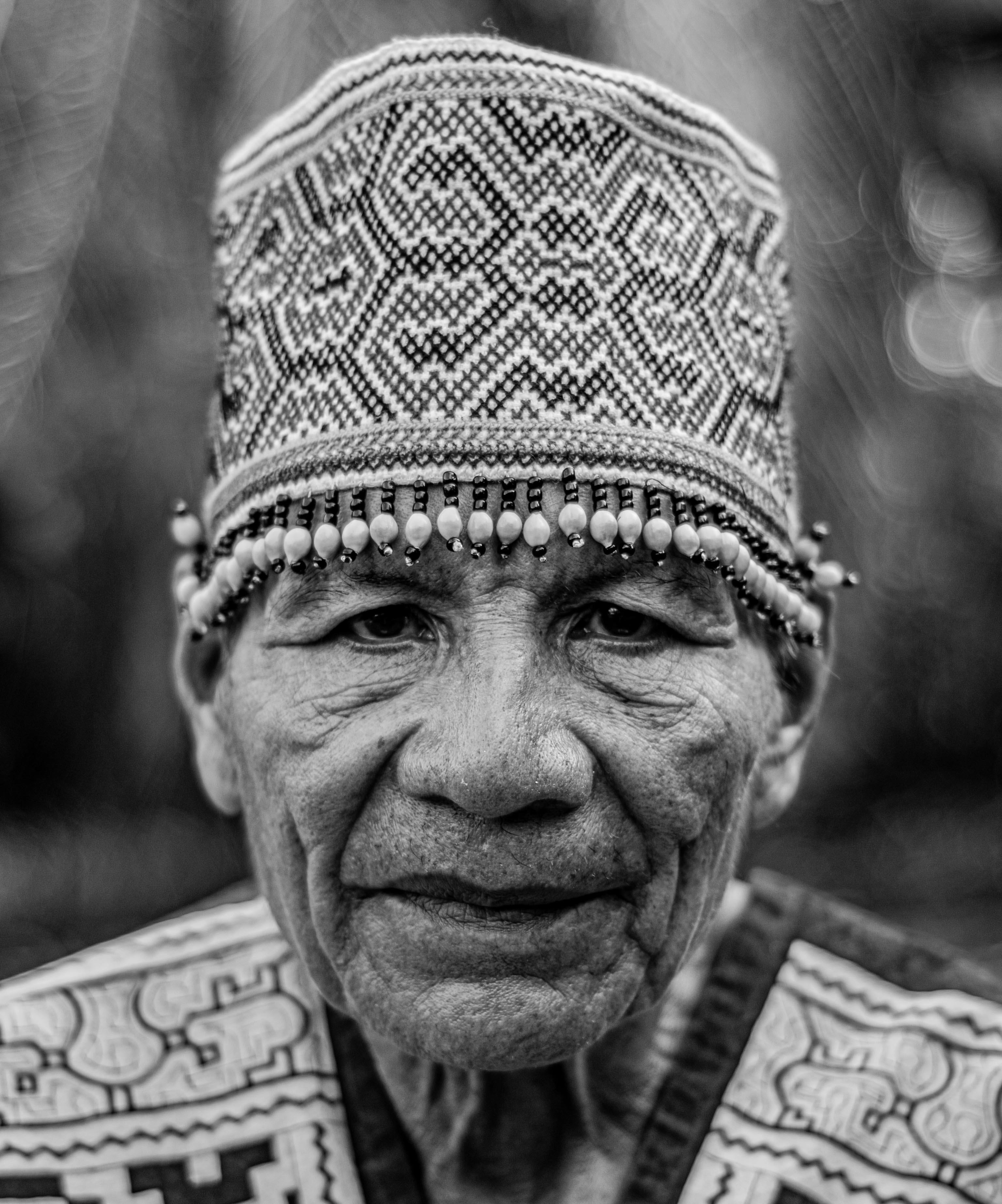 Black and white portrait of an elderly man in traditional tribal clothing and hat.