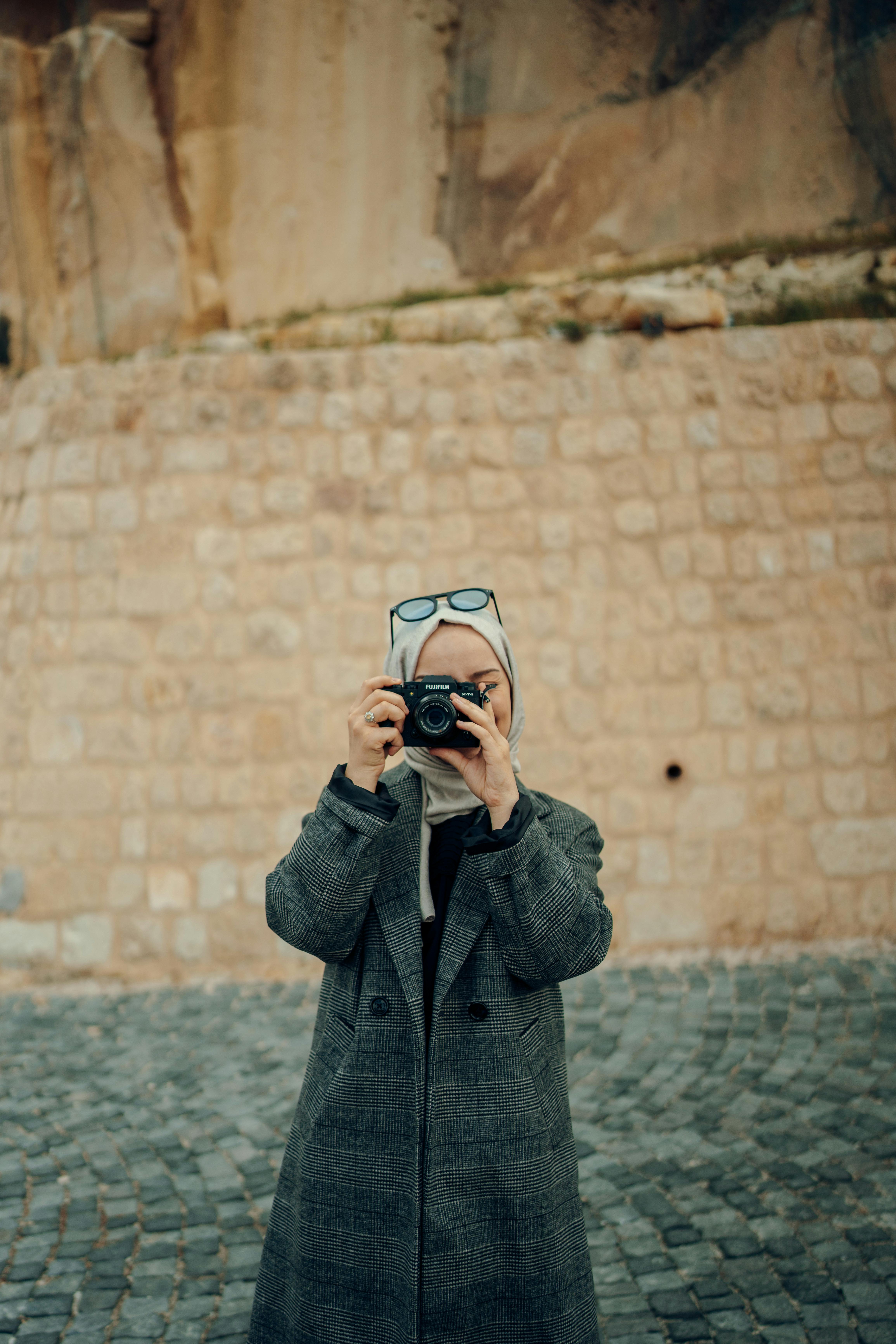 Woman in a hijab photographing with a camera against a stone wall.