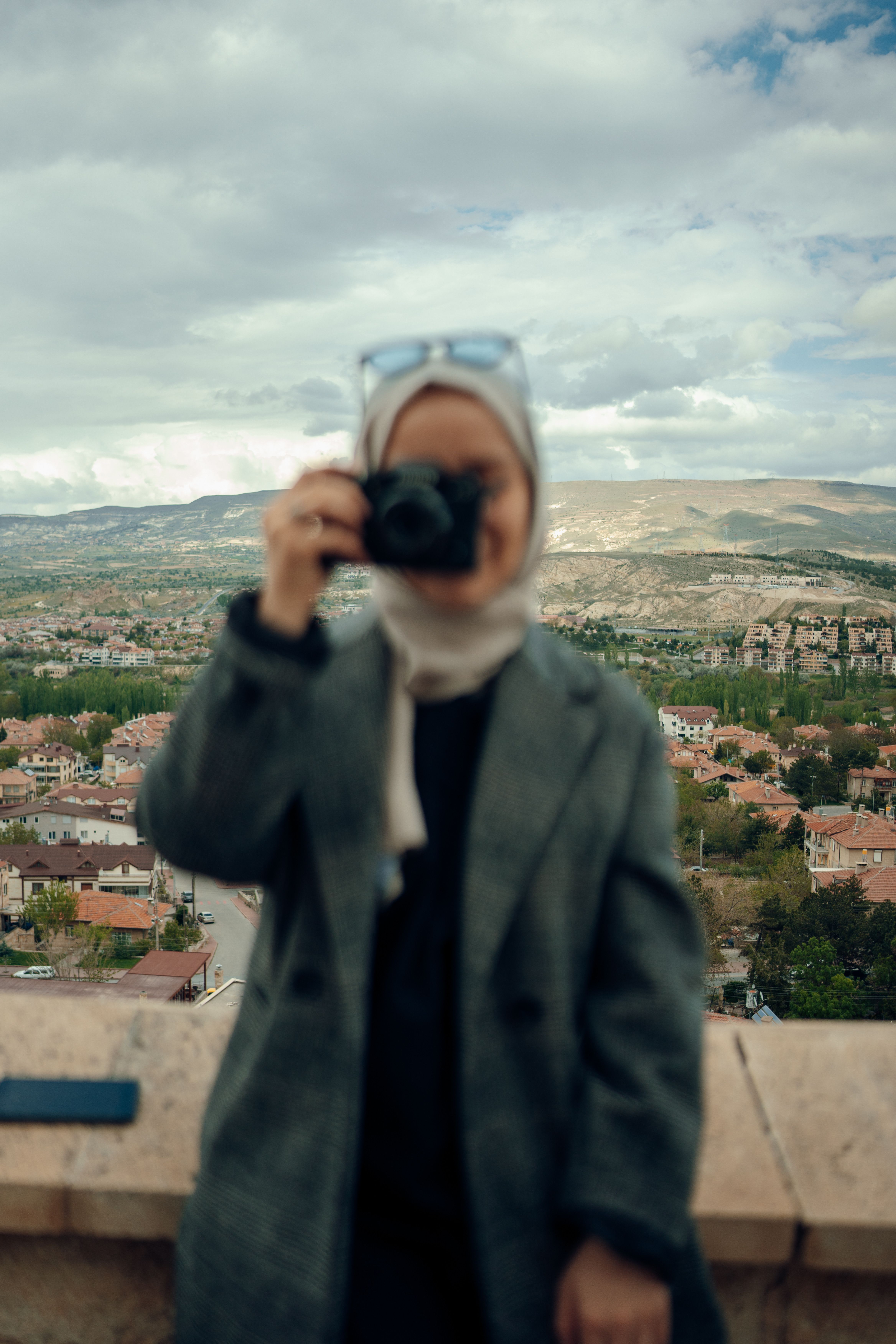 A blurred woman in a hijab captures an urban landscape with a camera.