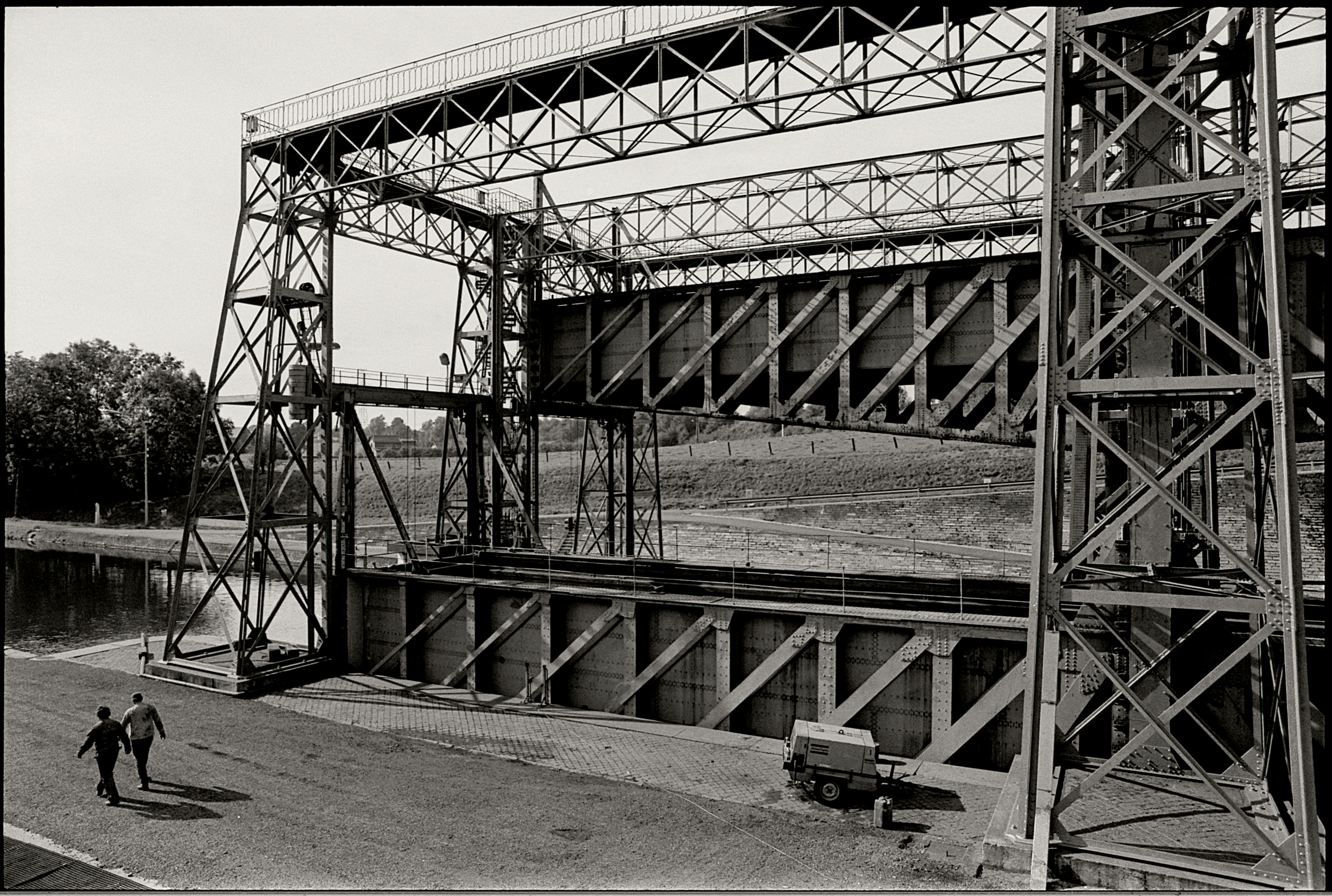 Black and white photo of a steel bridge over a canal, with two people walking nearby.