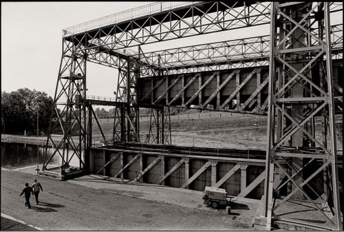 Free Black and white photo of a steel bridge over a canal, with two people walking nearby. Stock Photo