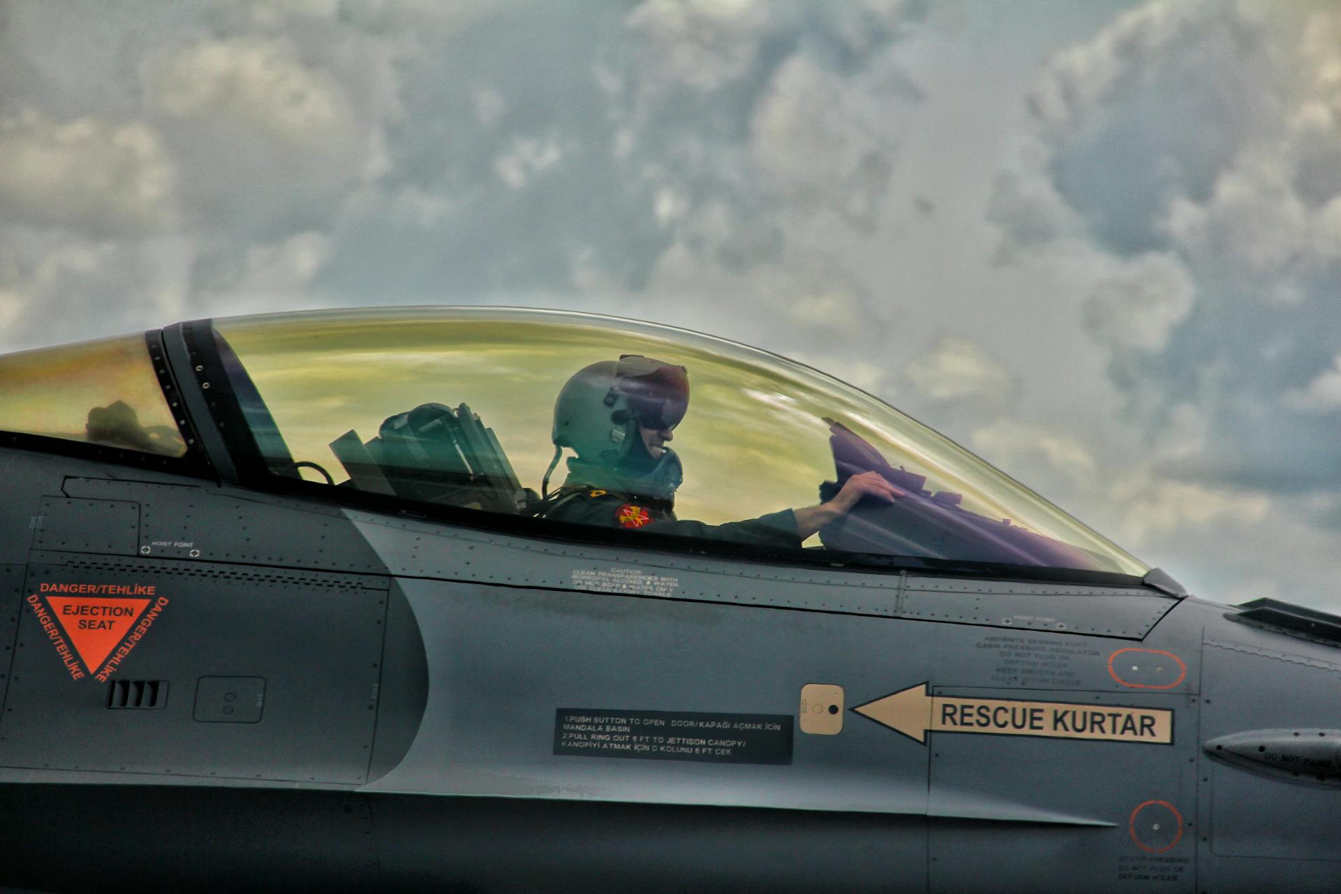 Close-up of a fighter jet pilot in cockpit, showcasing military aviation technology against clouded backdrop.