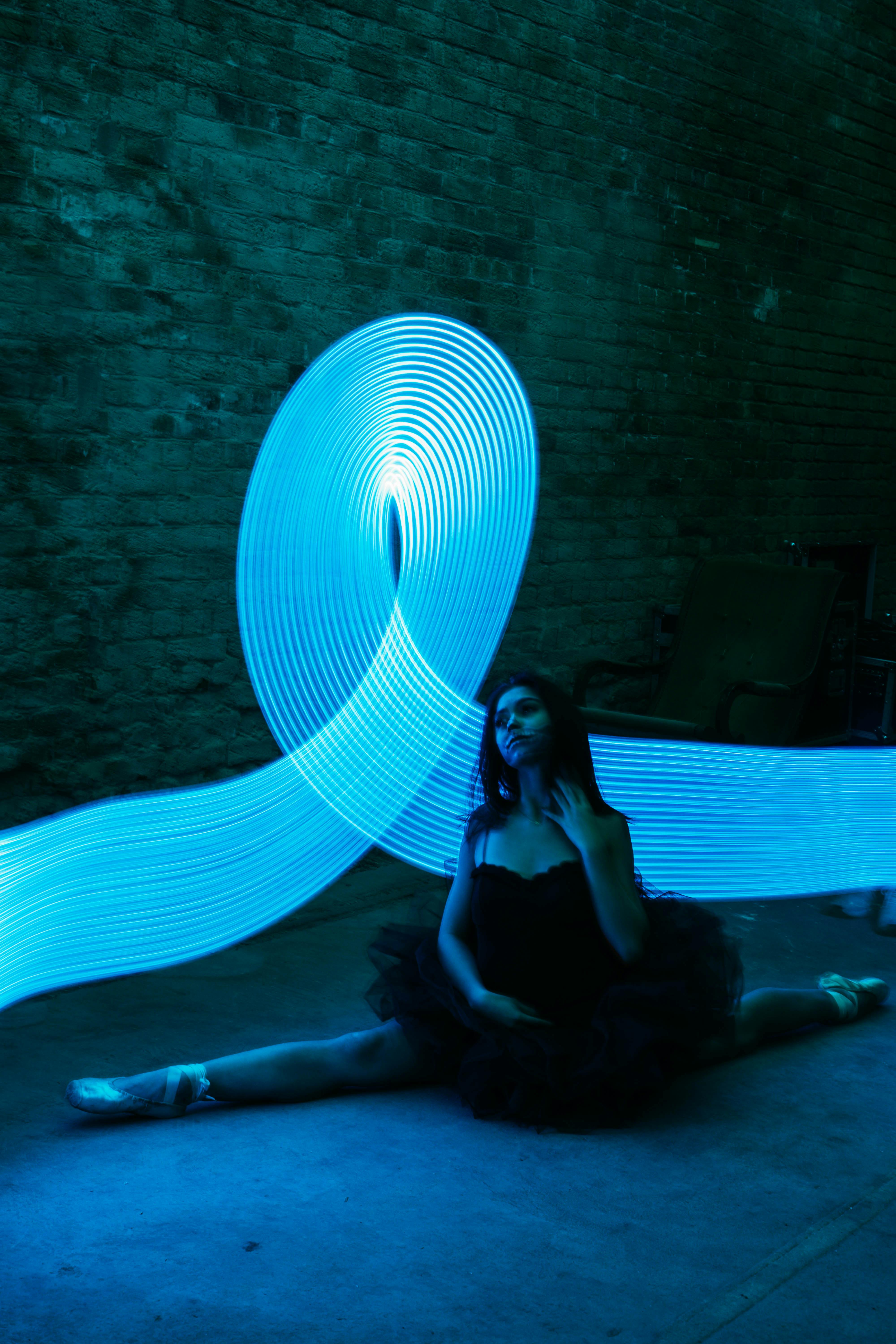 A ballerina in a split pose illuminated by blue neon light trails in a dark studio setting.