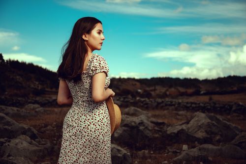 Free A woman in a floral dress holding a hat, stands in a rocky landscape under a vibrant sky. Stock Photo