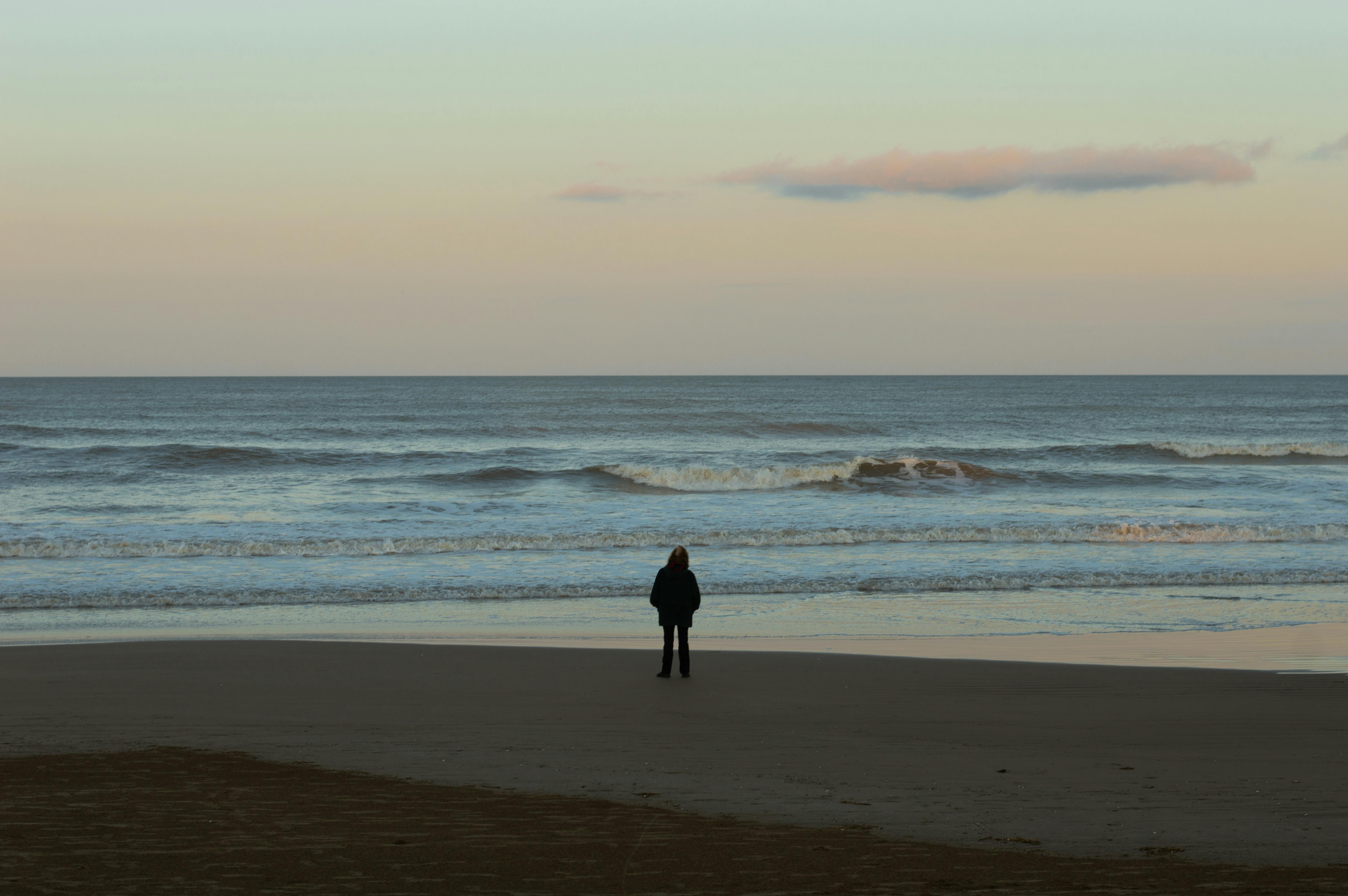 A person stands alone on Santa Teresita beach, gazing at the calm sea during twilight.