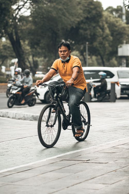Free A man in an orange shirt cycles on a sidewalk amidst city traffic, showcasing urban commuting. Stock Photo