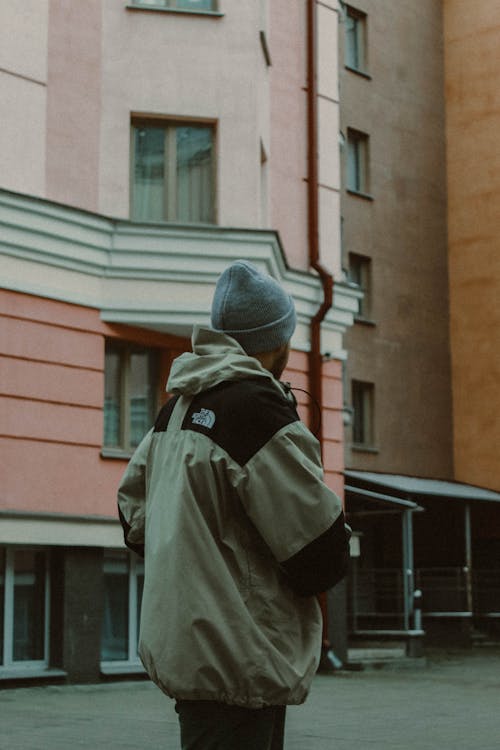 Young Man in a Winter Jacket and Hat Standing in front of a Residential Building in City 