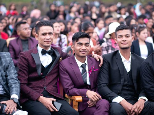 Free Three friends in suits sitting together, smiling at an outdoor event with a crowd backdrop. Stock Photo