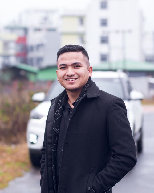 Free Smiling man in a black jacket stands confidently outdoors with a blurred urban background. Stock Photo