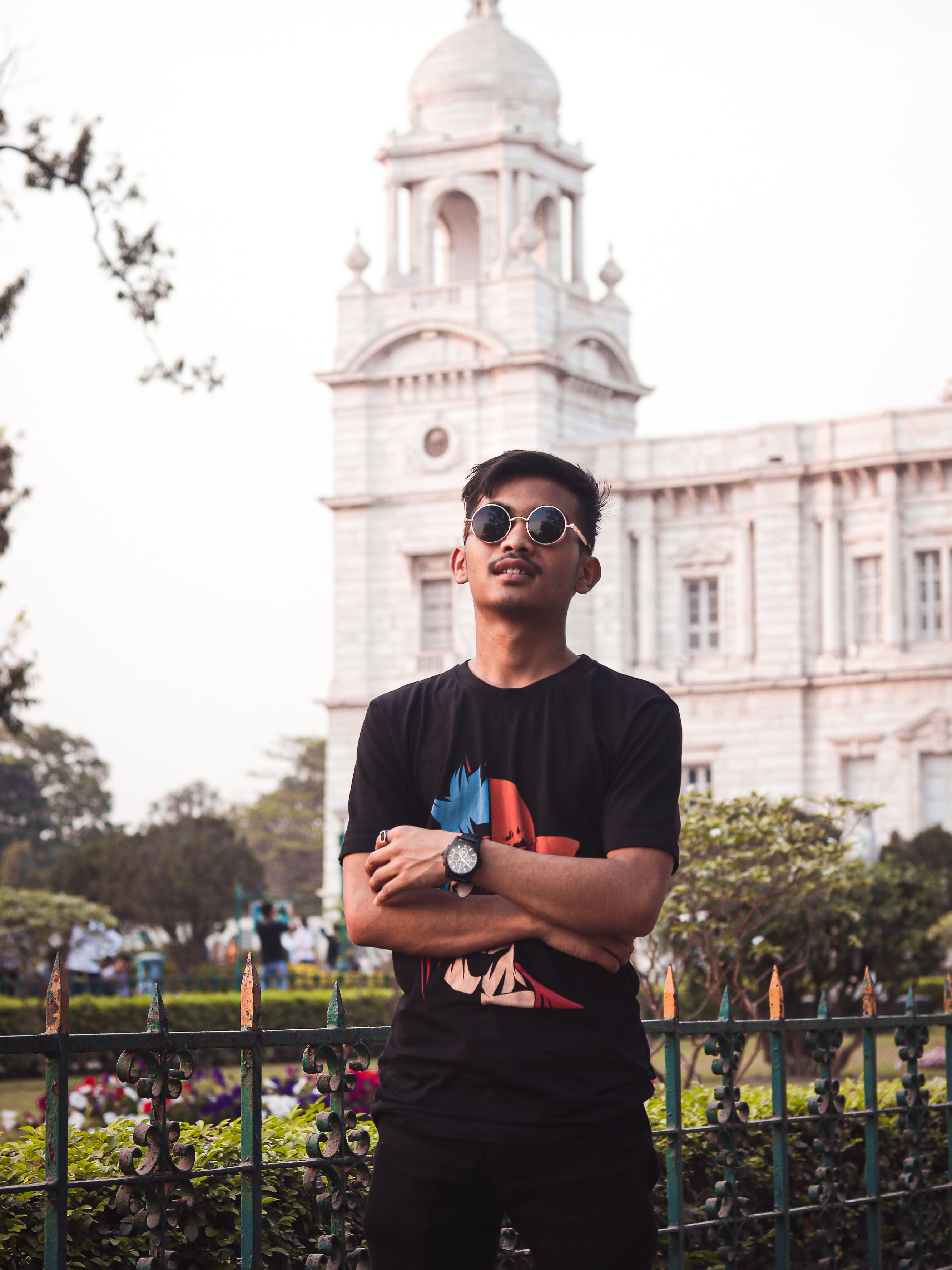 Fashionable young man with sunglasses in urban park setting, posing confidently before historic architecture.