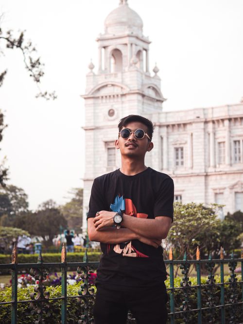 Free Fashionable young man with sunglasses in urban park setting, posing confidently before historic architecture. Stock Photo