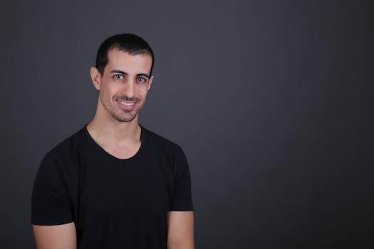 A confident young man in a black shirt smiles against a dark background, conveying friendliness and approachability.