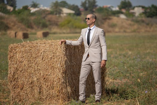 Confident businessman wearing sunglasses and a suit standing next to a hay bale in a rural field.