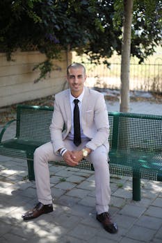 Confident businessman in a suit sitting on a green bench in an outdoor park setting.