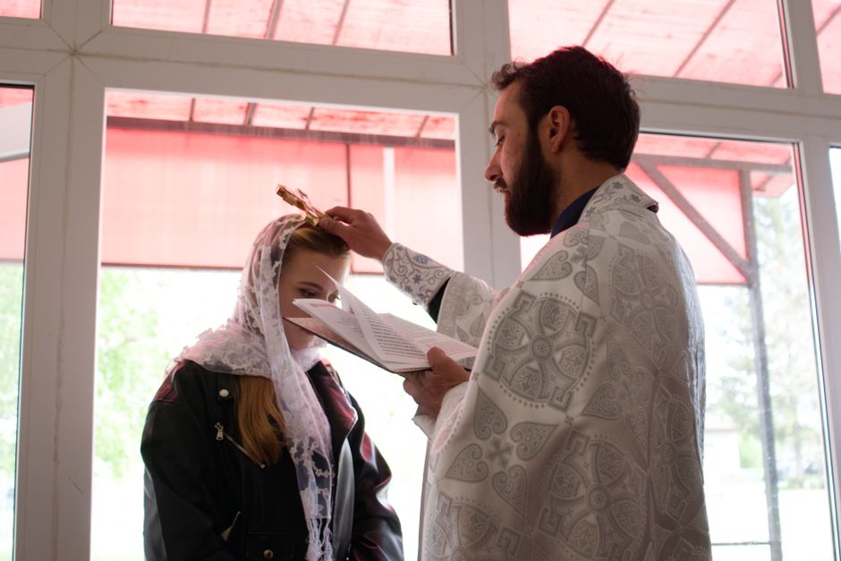 An Orthodox priest conducts a religious ceremony indoors, blessing a woman with a crown.