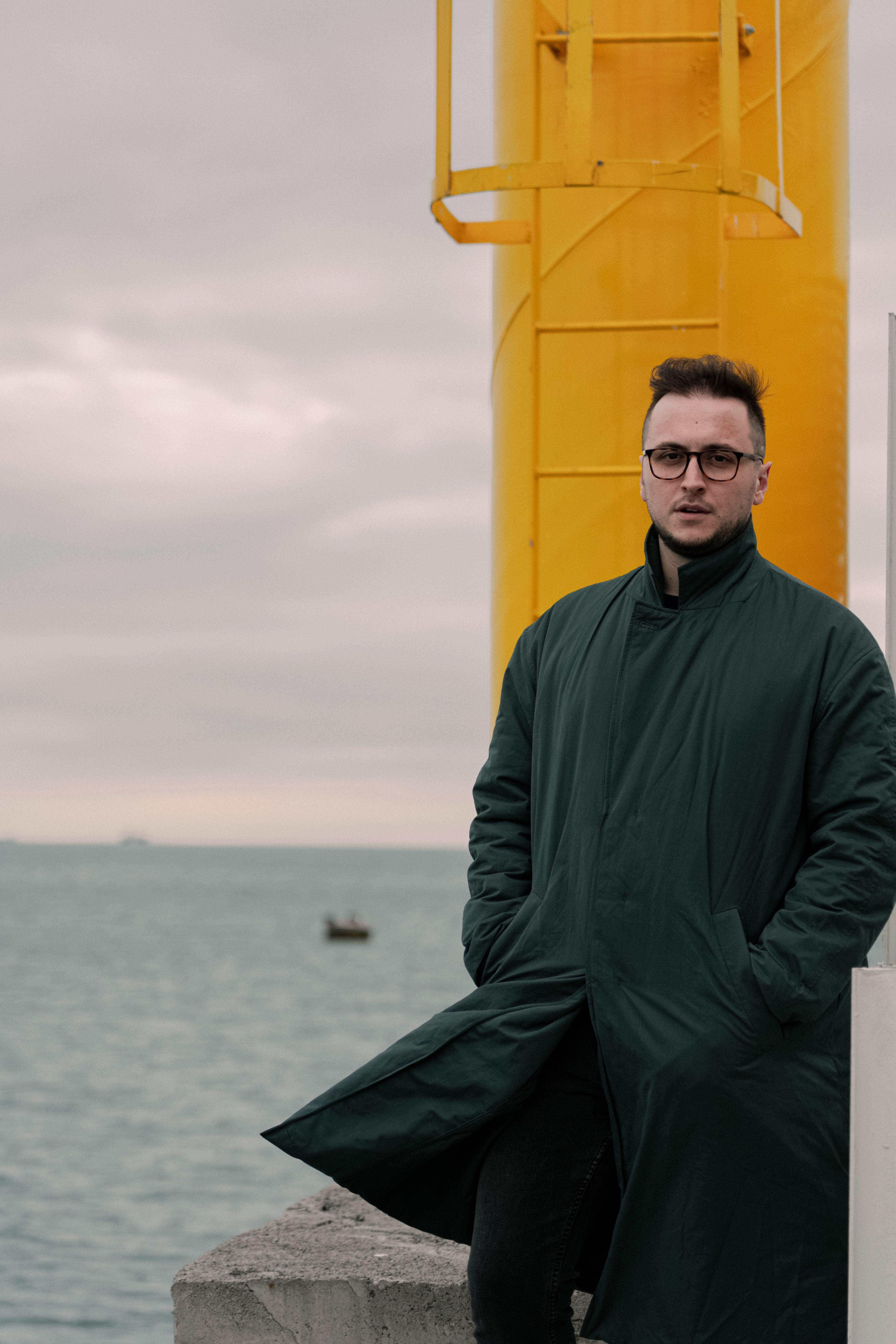 Man in coat posing by sea with yellow structure on an overcast day.