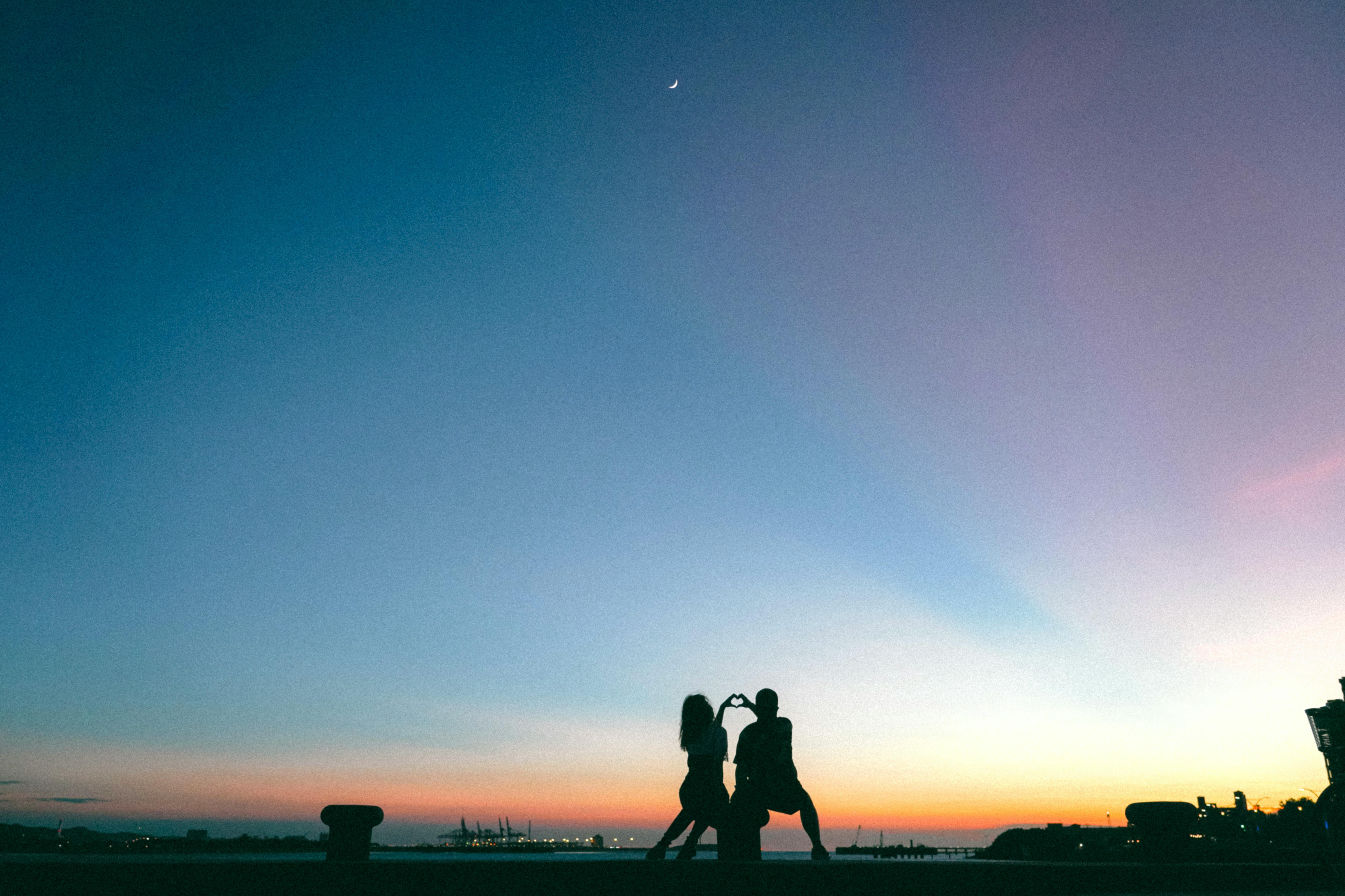 Couple creates heart shape with hands at sunset, city skyline in silhouette.