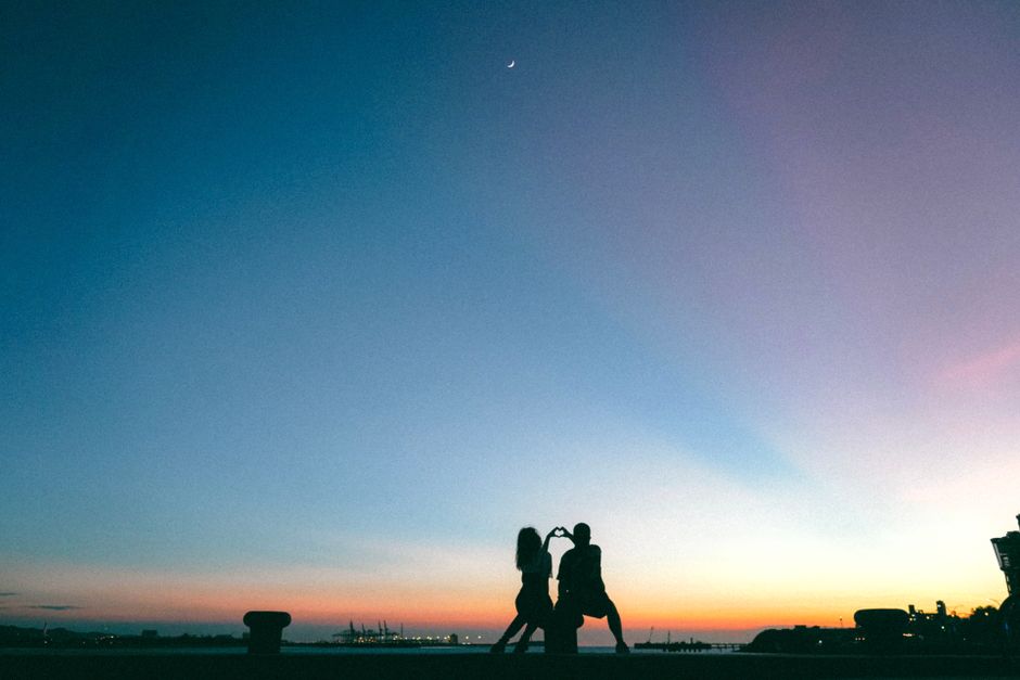 Couple creates heart shape with hands at sunset, city skyline in silhouette.