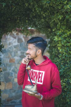 A young man in a red hoodie holding a vintage telephone in a lush Srinagar garden.