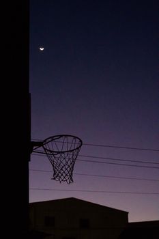 Silhouette of a basketball hoop against a twilight sky with a crescent moon.
