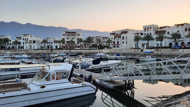 Marina with motorboats and traditional buildings in As Sifah, Oman at sunset.