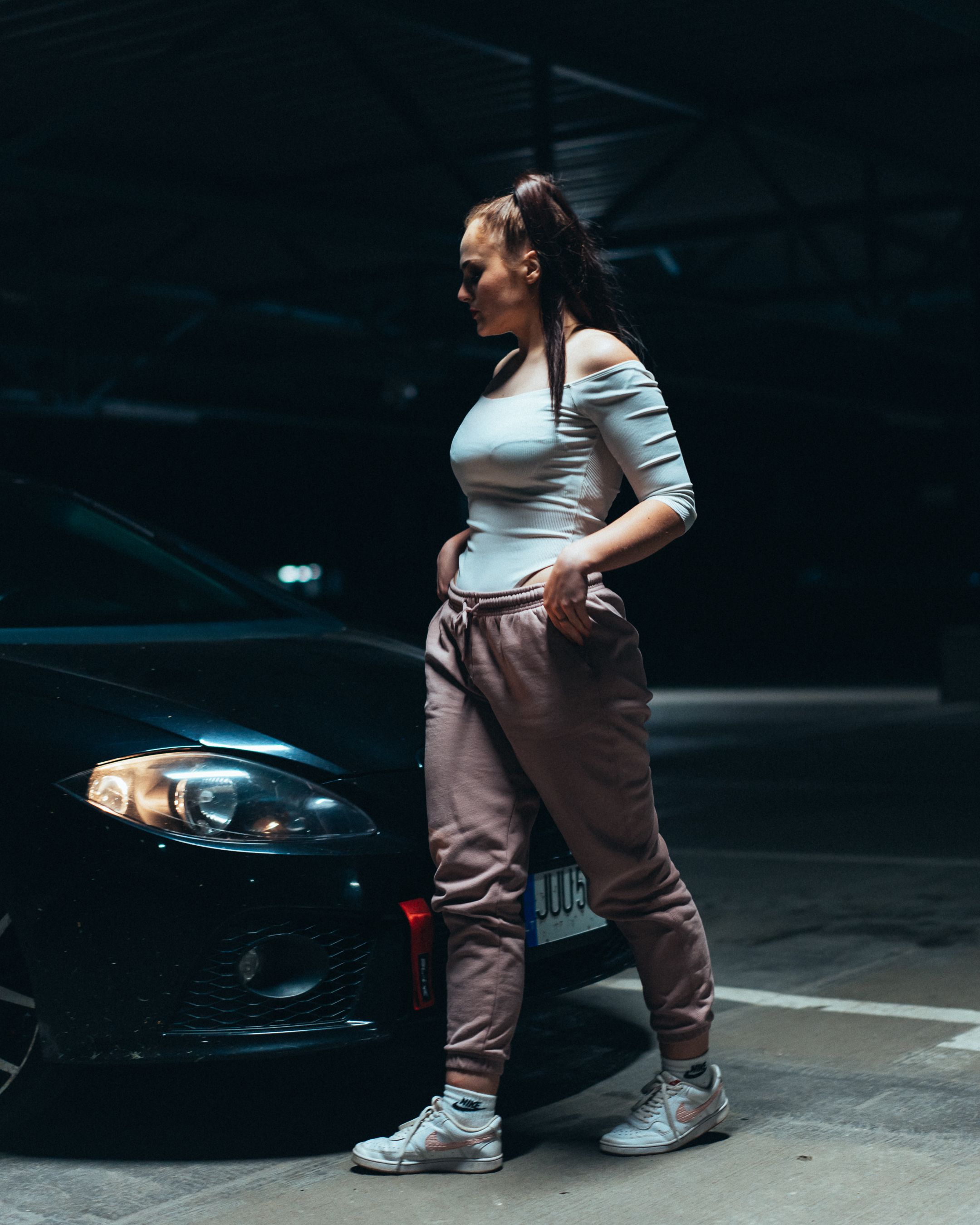 A fashionable woman poses confidently beside a car in an urban parking lot at night.