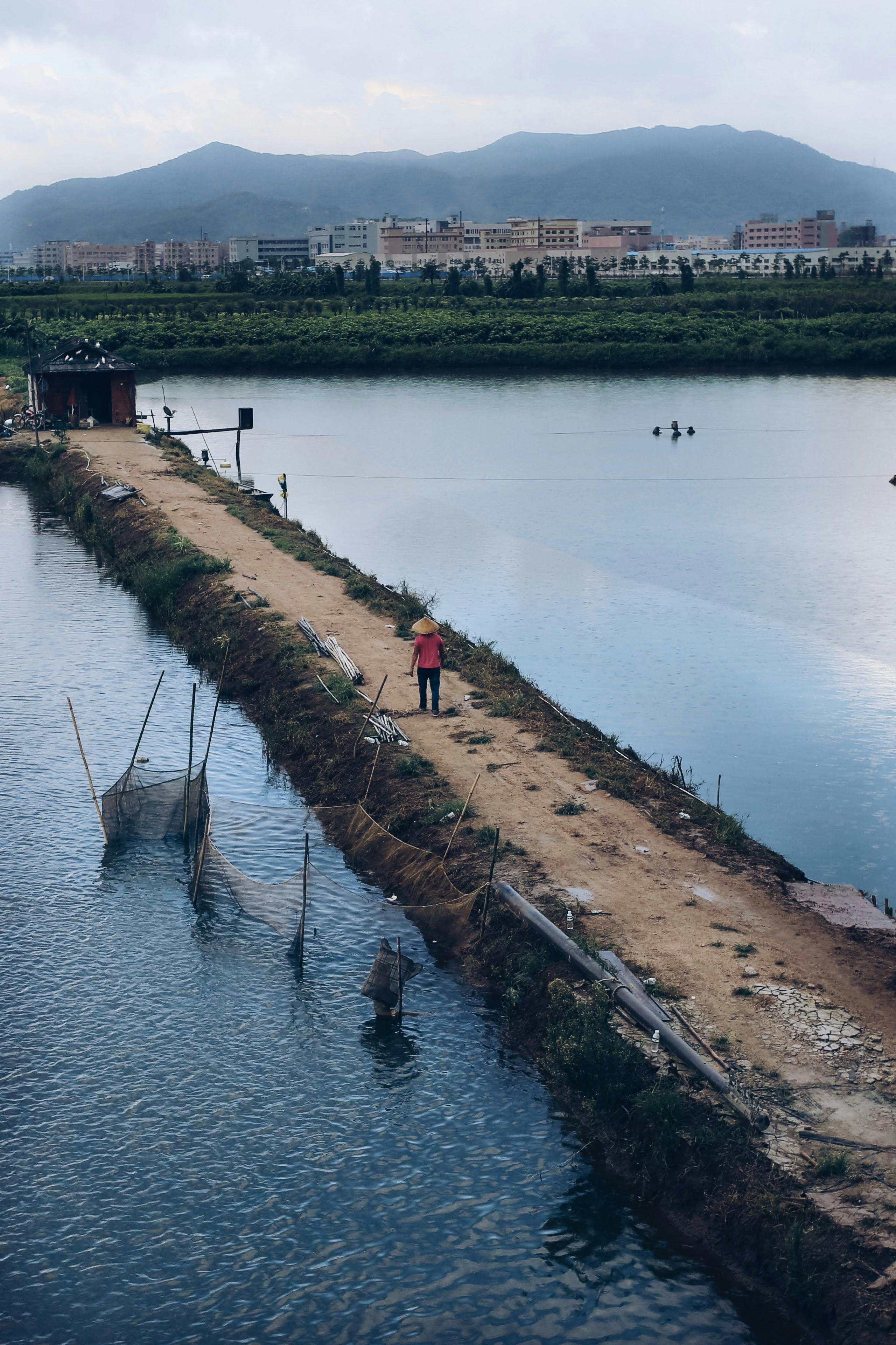 A fisherman walks along a narrow path by a lake, framed by mountains and a distant town.